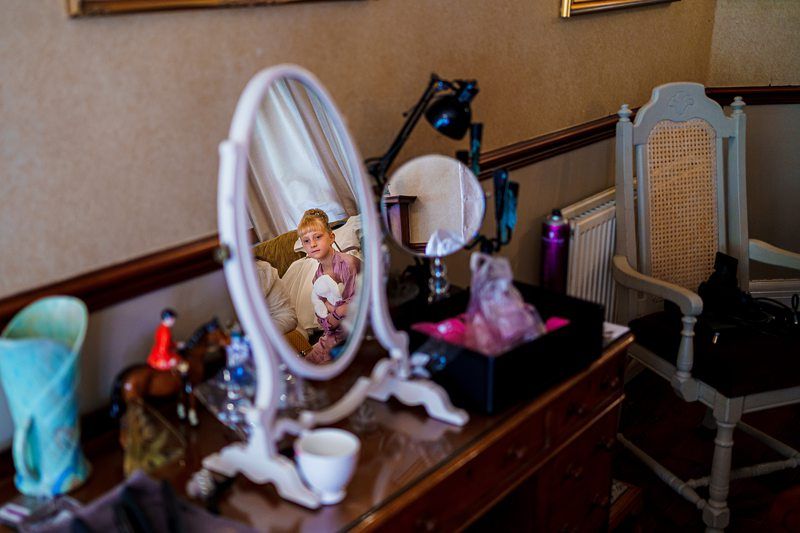 A young child and an adult are seen sitting on a couch, visible through a mirror on a vanity table. The vanity table has various items including a lamp, makeup accessories, and a cup. The room has warm lighting and classic furniture. Image by Documentary Wedding Photographer Lewis Bishop Photography.