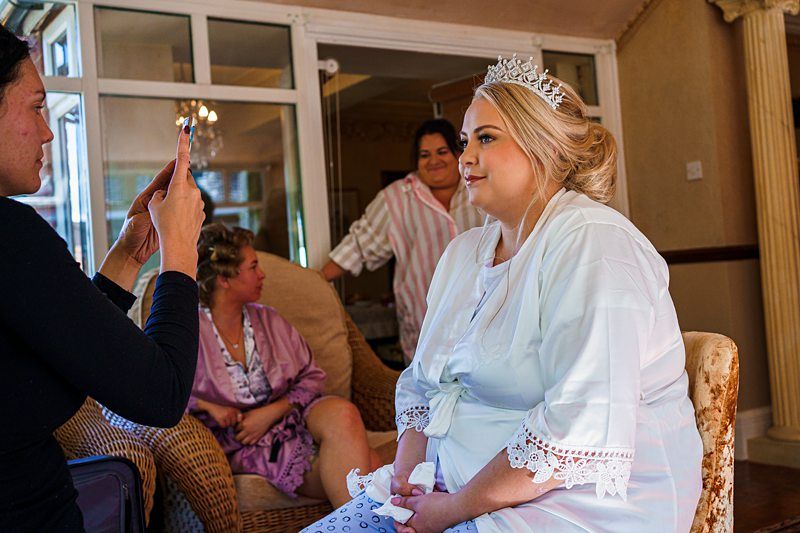 A bride in a white robe and tiara sits in a wicker chair while someone takes a picture of her using a phone. Two other women, one in a mauve robe and another in striped pajamas, are in the background chatting and smiling inside a cozy room. Image by Documentary Wedding Photographer Lewis Bishop Photography.