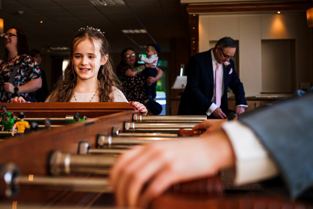 A young girl with long brown hair smiles while playing foosball. In the background, adults, including a man in a suit and a woman with a baby, converse and watch. The setting appears to be a well-lit indoor event or party. Image by Documentary Wedding Photographer Lewis Bishop Photography.