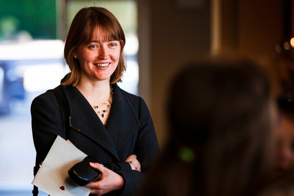 A woman with shoulder-length brown hair is smiling and standing indoors, holding a white envelope and a black object. She is wearing a dark coat and a necklace. The background is blurred and includes another person in the foreground. Image by Documentary Wedding Photographer Lewis Bishop Photography.
