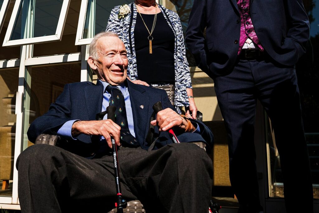 An elderly man wearing a dark suit and patterned tie smiles while seated outdoors, holding a cane. A woman and another man, also dressed formally, stand behind him. Sunlight illuminates the group, with a building featuring large windows in the background. Image by Documentary Wedding Photographer Lewis Bishop Photography.