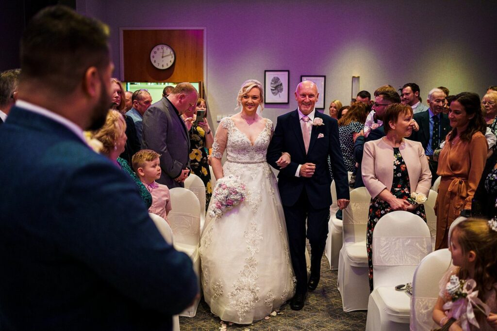 A bride, in a white wedding dress and holding a bouquet, walks down the aisle accompanied by an older man wearing a navy suit. They are surrounded by smiling guests seated on both sides, all dressed in formal attire. A clock and framed pictures are visible on the wall. Image by Documentary Wedding Photographer Lewis Bishop Photography.