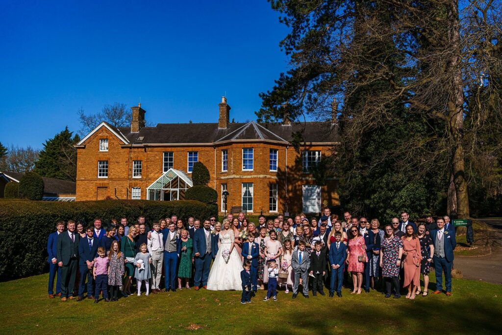A large group of people, dressed in formal attire, pose in front of a stately brick building with manicured gardens. The central focus is a bride and groom surrounded by family and friends, all smiling for the camera under a clear blue sky. Image by Documentary Wedding Photographer Lewis Bishop Photography.