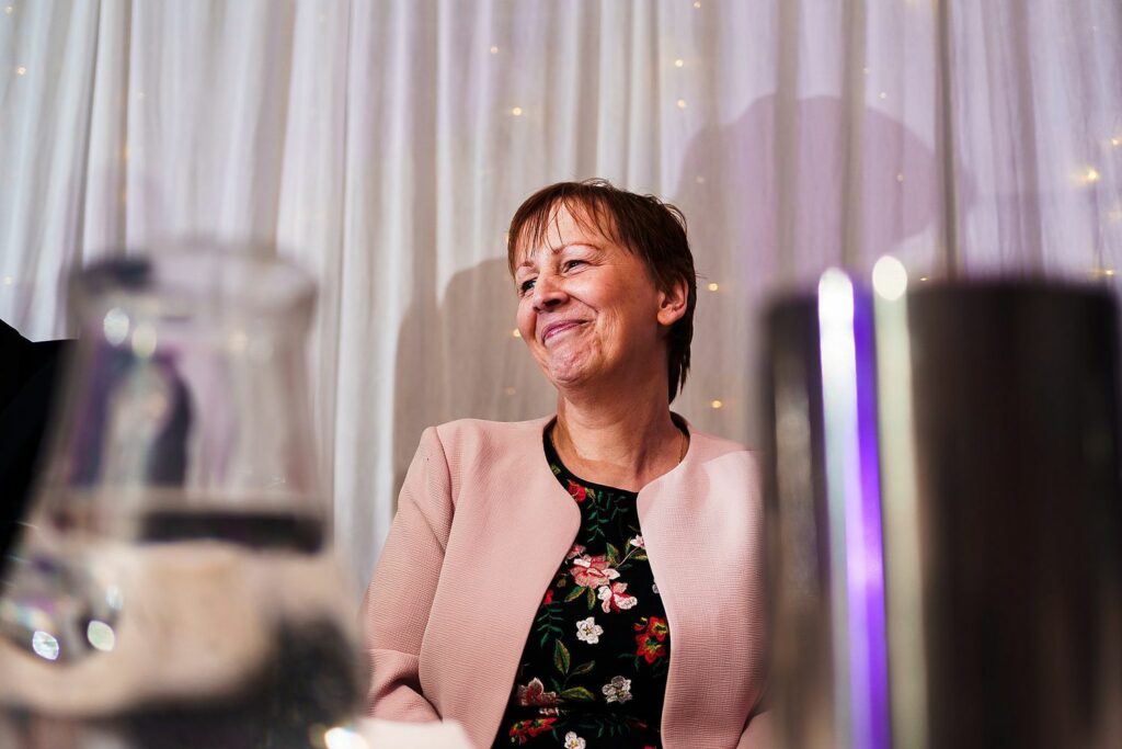 A woman with short hair is smiling while seated in front of a curtain with small lights. She is wearing a floral-patterned top and a light pink blazer. Various out-of-focus glassware is in the foreground. Image by Documentary Wedding Photographer Lewis Bishop Photography.