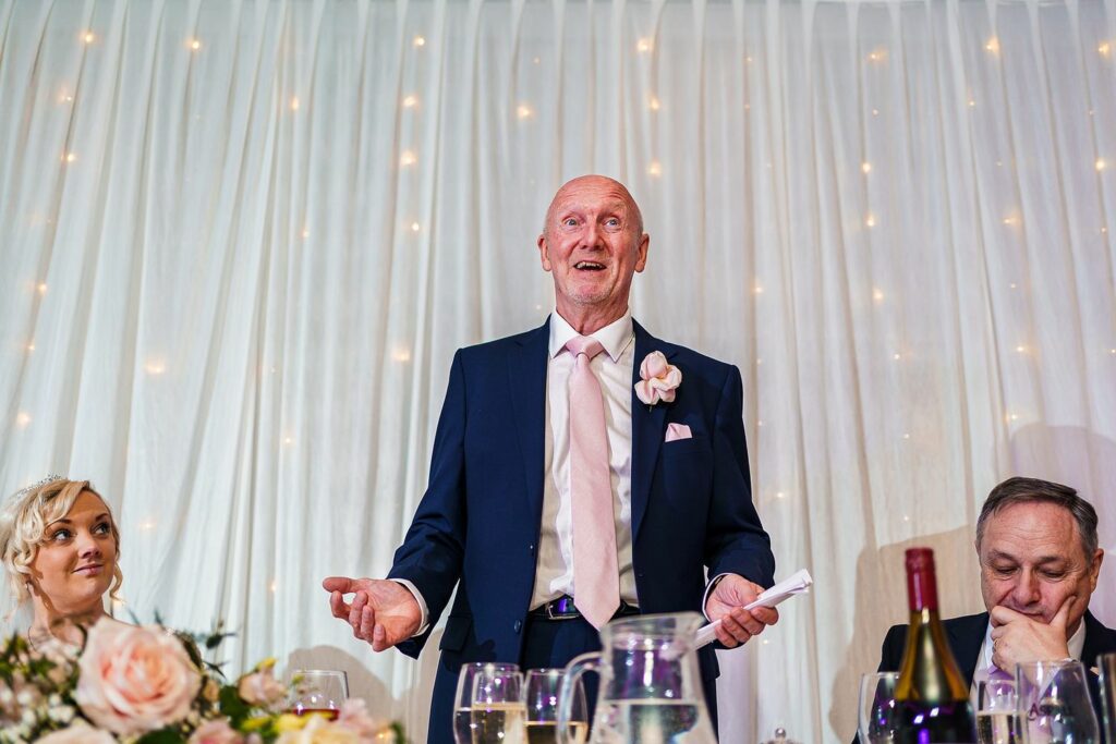 An older man in a navy suit and light pink tie stands giving a speech, smiling, with a glass in one hand and notes in the other. A younger woman and a middle-aged man sit nearby. The backdrop is illuminated with warm string lights. Image by Documentary Wedding Photographer Lewis Bishop Photography.