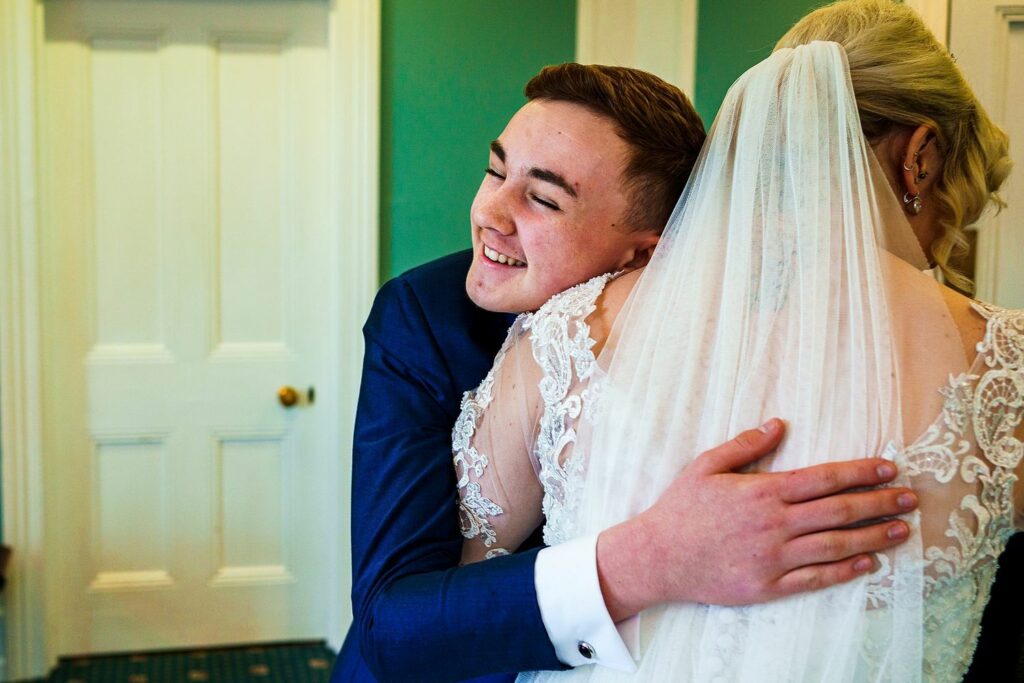 A young man in a blue suit embraces a bride wearing a lace wedding dress and veil. They are standing in a warmly lit room with green walls and white trim. The man is smiling joyfully with his eyes closed. Image by Documentary Wedding Photographer Lewis Bishop Photography.