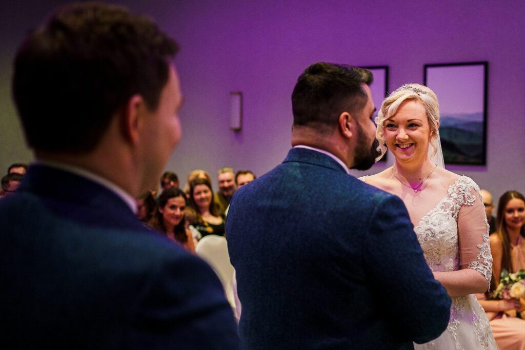A bride and groom stand facing each other, smiling, during their wedding ceremony. The bride is wearing a white lace gown and veil, while the groom is in a dark blue suit. Guests are seated in the background, attentively watching the couple. Image by Documentary Wedding Photographer Lewis Bishop Photography.