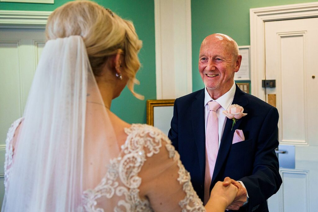 A bride with a lace dress and veil holds hands with an older man wearing a suit and a pink tie. Both are smiling warmly at each other in a room with green walls and white trim. The interaction suggests a heartfelt and joyous moment. Image by Documentary Wedding Photographer Lewis Bishop Photography.