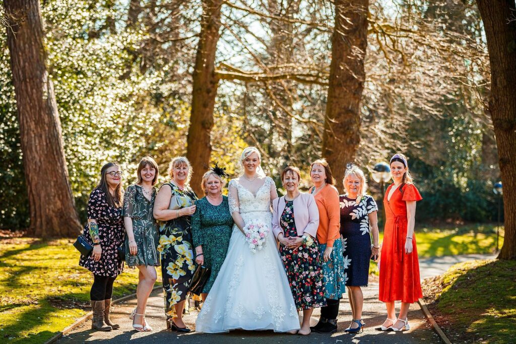A bride in a white gown stands outdoors with a group of ten women, all dressed in colorful dresses and smiling for the photo. They are in a wooded area with tall trees and sunlight filtering through. The background suggests it is a clear, sunny day. Image by Documentary Wedding Photographer Lewis Bishop Photography.