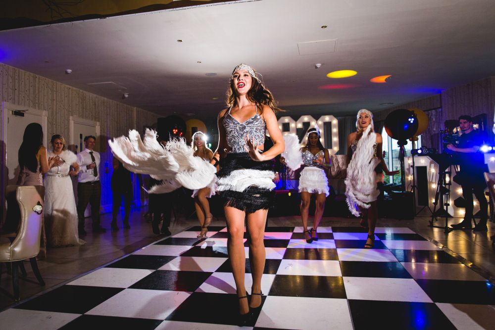 A group of dancers in 1920s flapper-style costumes perform on a black and white checkered dance floor. The lead dancer in front wears a silver sequined top, black fringe skirt, and headband with a feather, while others hold white feathered fans. Image by Documentary Wedding Photographer Lewis Bishop Photography.