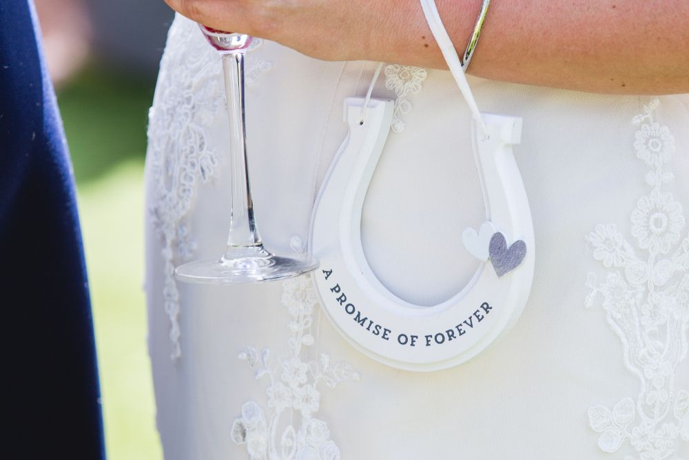 A close-up of a bride holding a glass of champagne in her right hand. She is wearing a white lace wedding dress, and her left arm holds a white horseshoe with the text "A PROMISE OF FOREVER" and two small heart embellishments. Image by Documentary Wedding Photographer Lewis Bishop Photography.