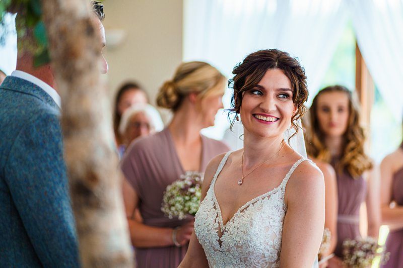 A bride with brown curly hair, wearing a white lace wedding dress, smiles joyfully during a ceremony. She is surrounded by bridesmaids in matching mauve dresses, holding bouquets, while the groom, slightly out of frame, stands beside her. Image by Documentary Wedding Photographer Lewis Bishop Photography.