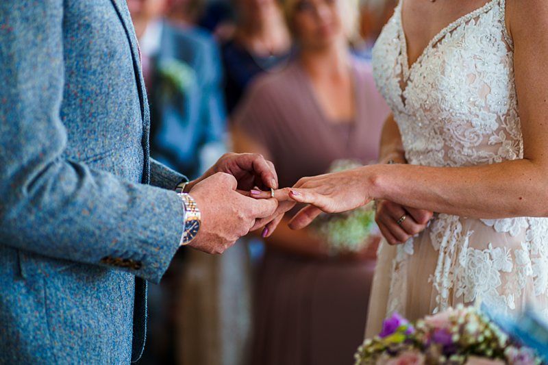 A groom in a blue suit places a ring on the bride's finger at a wedding ceremony. The bride is wearing a white lace gown and holding a bouquet of flowers. They are surrounded by blurred guests in the background. Image by Documentary Wedding Photographer Lewis Bishop Photography.