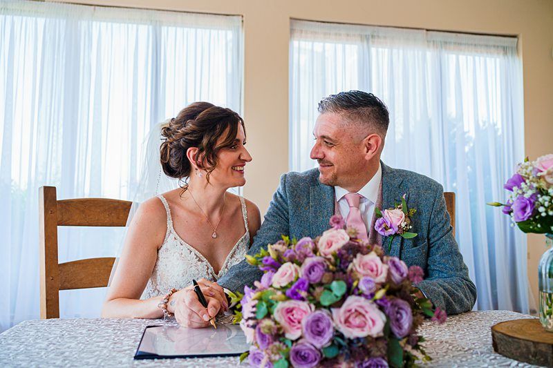 A bride and groom sit at a table adorned with a lace tablecloth and a bouquet of purple and pink roses. They are smiling and looking into each other's eyes as the bride holds a pen, possibly signing a document. Two large windows with white curtains are in the background. Image by Documentary Wedding Photographer Lewis Bishop Photography.