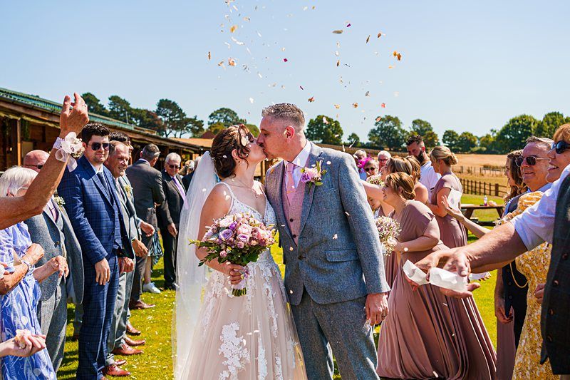 A newlywed couple shares a kiss outdoors while guests toss confetti around them. The bride holds a bouquet of flowers and wears a lace wedding gown with a veil. The groom, dressed in a grey suit, lovingly holds her hand. Guests, dressed in colorful attire, applaud and smile. Image by Documentary Wedding Photographer Lewis Bishop Photography.