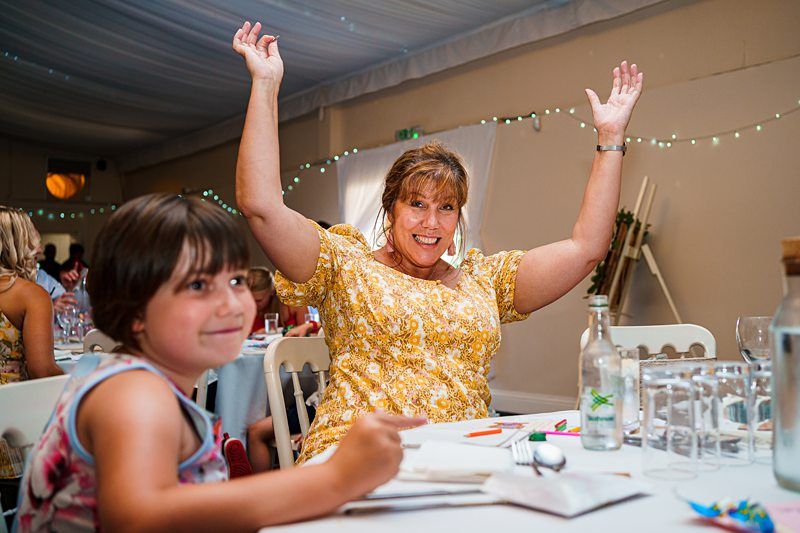 A woman in a yellow floral dress is raising her arms and smiling joyfully while seated at a table set for a meal. A young child sits beside her, focused on coloring. The room is decorated with string lights, and the table has various items, including glasses and a water bottle. Image by Documentary Wedding Photographer Lewis Bishop Photography.