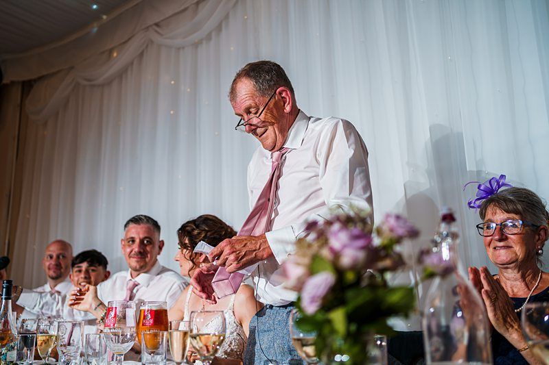 A man in a light pink tie and glasses stands and speaks while holding notes at a wedding reception. Others seated at the table, including a woman with a purple fascinator, listen attentively. Glasses and flowers are seen in the foreground. Image by Documentary Wedding Photographer Lewis Bishop Photography.