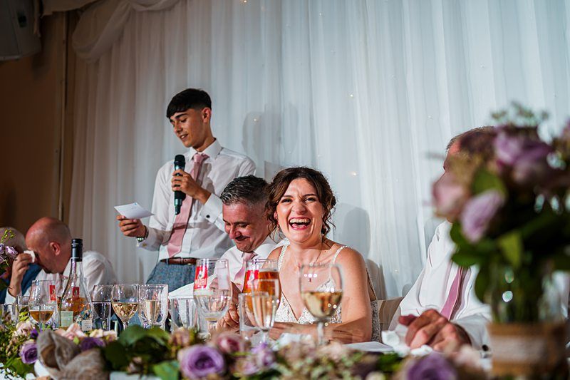 A wedding reception scene where a groom gives a speech while holding a piece of paper. The bride, seated next to him, is joyfully laughing. Other guests are also seated at the table, which is decorated with flowers and glassware. Image by Documentary Wedding Photographer Lewis Bishop Photography.