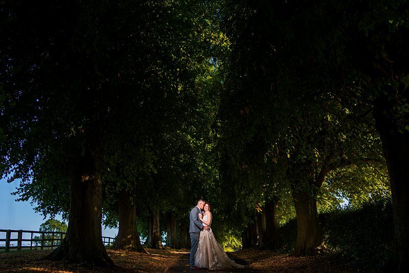 A couple in wedding attire stands closely together in a tender embrace under a canopy of large, leafy trees. The sun casts a soft light through the branches, creating a romantic, secluded atmosphere. The surrounding area is shaded and serene. Image by Documentary Wedding Photographer Lewis Bishop Photography.