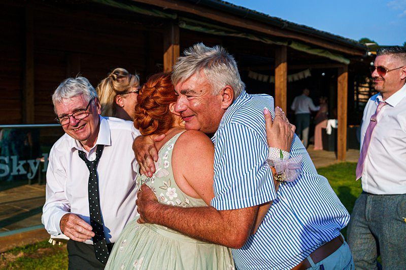A group of people enjoys a moment at an outdoor event. A man in a blue striped shirt is embracing a woman in a light green dress. Two men stand nearby, one in a white shirt and tie, both smiling warmly. The background features a wooden structure and clear skies. Image by Documentary Wedding Photographer Lewis Bishop Photography.