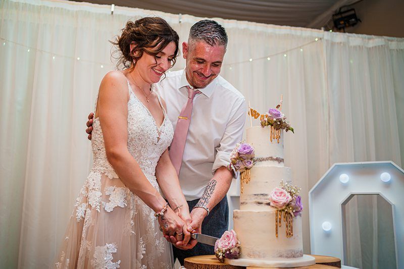 A couple wearing wedding attire cuts into a tiered wedding cake adorned with pink and purple flowers. The bride wears a white lace gown, and the groom is dressed in a white shirt with a pink tie. They stand in front of a white curtain backdrop. Image by Documentary Wedding Photographer Lewis Bishop Photography.