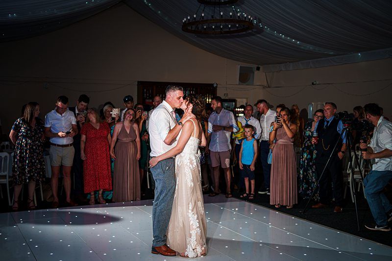 A bride and groom share their first dance on a white, illuminated dance floor. Guests surround them, some capturing the moment with phones and cameras. The venue is decorated with draped fabric and string lights, creating a romantic atmosphere. Image by Documentary Wedding Photographer Lewis Bishop Photography.