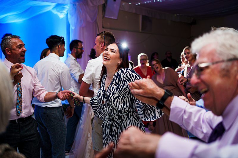 A group of people dancing joyfully in a dimly-lit room with blue lighting. The woman in the center wearing a zebra-print dress is smiling widely. The atmosphere is lively and festive, with hands joined and expressions of happiness all around. Image by Documentary Wedding Photographer Lewis Bishop Photography.