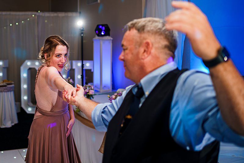 A young woman in a pink dress dances with an older man in a blue shirt and black vest at an indoor event. They hold hands and spin together, surrounded by soft lighting and decor, including a large "LOVE" sign in the background. Image by Documentary Wedding Photographer Lewis Bishop Photography.