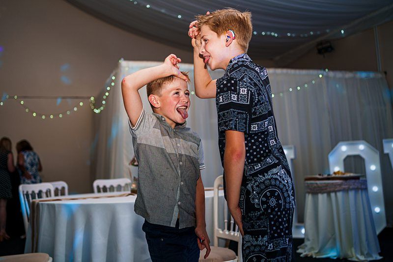 Two boys are playing and being silly together in a decorated room. The older boy, wearing a patterned outfit, is placing his hand on the younger boy’s head, who sticks his tongue out playfully. The background has chairs, tables, and twinkling lights. Image by Documentary Wedding Photographer Lewis Bishop Photography.