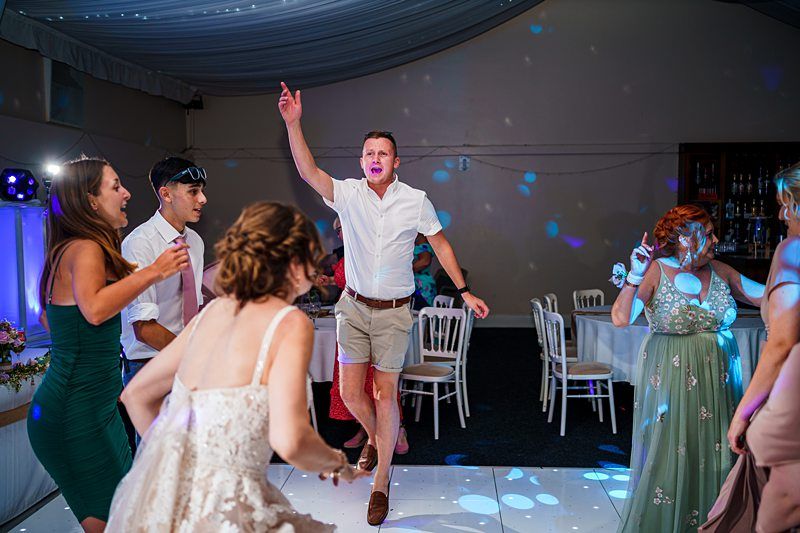 A group of people are dancing energetically at an indoor event. A man in a white shirt and shorts is jumping with one arm raised. The women around him are dressed in dresses, one in a wedding gown. The scene is vibrant with colorful lights and festive decorations. Image by Documentary Wedding Photographer Lewis Bishop Photography.