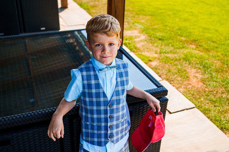 A young boy with short blonde hair, dressed in a blue and grey plaid vest, blue shirt, and bow tie, stands outdoors on a patio. He holds a red baseball cap in his right hand and smiles at the camera. A grassy yard is visible in the background. Image by Documentary Wedding Photographer Lewis Bishop Photography.