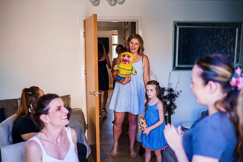 A woman holding a baby enters a room with a smile, joined by other women and a young girl. They are all enjoying a cheerful moment in a warmly decorated living space, suggesting a family gathering or celebration. Image by Documentary Wedding Photographer Lewis Bishop Photography.
