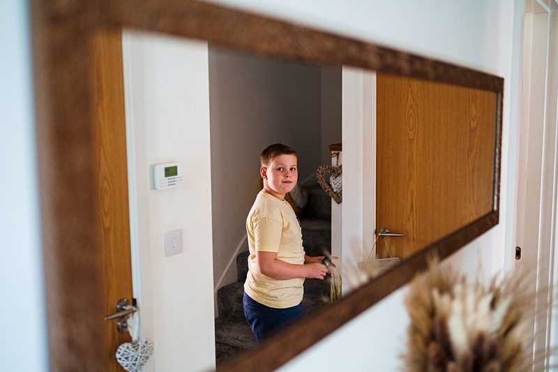 A boy in a yellow t-shirt stands in a hallway, looking back towards the camera. He is holding a game controller and is reflected in a large mirror hanging on the wall. The hallway is decorated with neutral colors and wooden doors. Image by Documentary Wedding Photographer Lewis Bishop Photography.