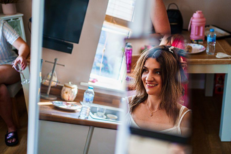 A woman with shoulder-length hair smiles as she looks into a mirror. Behind her, a person helps style her hair. The room has a cozy atmosphere with a small table, a pink container, a plate, a water bottle, and a TV in the background. Image by Documentary Wedding Photographer Lewis Bishop Photography.