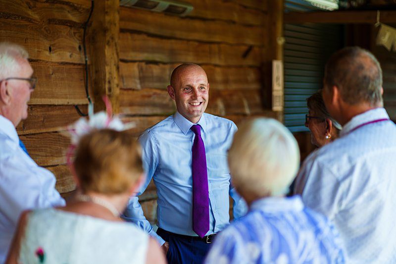 A man in a blue shirt and purple tie stands and smiles while talking to a group of people. The setting appears to be informal, with rustic wooden walls in the background. The people in the group are facing the man, listening attentively. Image by Documentary Wedding Photographer Lewis Bishop Photography.