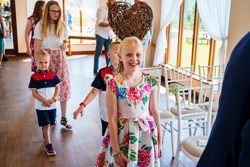 A group of children dressed in vibrant outfits walk down the aisle in a well-lit room with large windows. The girl in front wears a colorful floral dress and smiles, followed by other boys and girls in casual summer attire. A decorative heart made of vines is visible. Image by Documentary Wedding Photographer Lewis Bishop Photography.