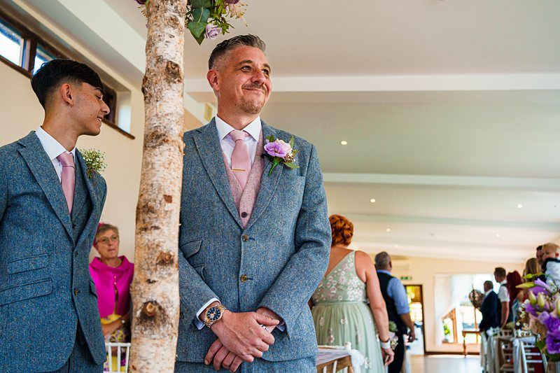 A man in a blue suit with a pink tie and a purple boutonniere stands smiling near a birch tree, next to a younger man also in a blue suit and pink tie. People are seated and standing in a bright room, suggesting a wedding ceremony. Image by Documentary Wedding Photographer Lewis Bishop Photography.