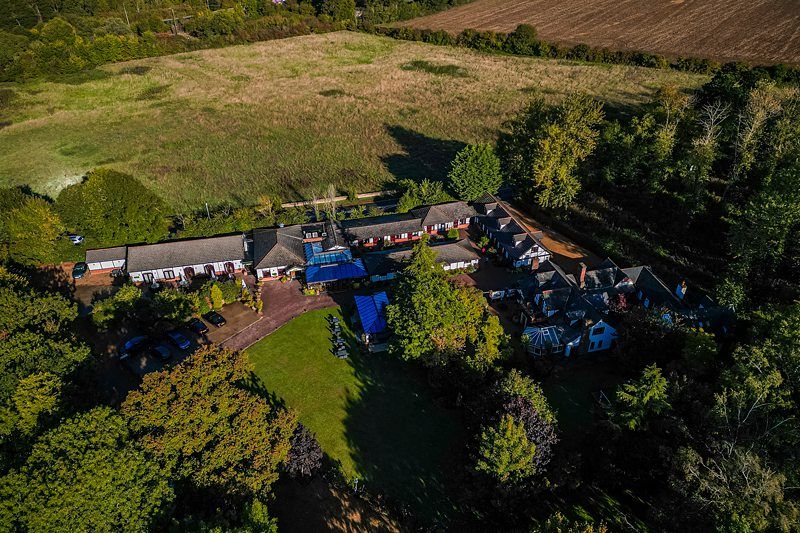 Aerial view of a large countryside property surrounded by trees and fields. The estate includes multiple buildings with red roofs, connected by pathways and open spaces with greenery. There are parked cars and blue outdoor coverings visible in the central area. Image by Documentary Wedding Photographer Lewis Bishop Photography.