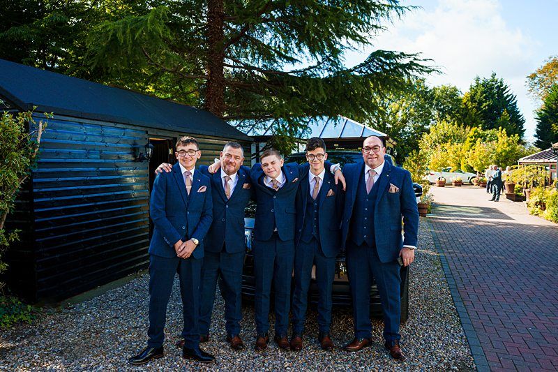 Five men dressed in blue suits and pink ties stand together, smiling, outside on a sunny day. They are on a gravel path next to a black building. Trees and landscaped gardens are visible in the background. Image by Documentary Wedding Photographer Lewis Bishop Photography.