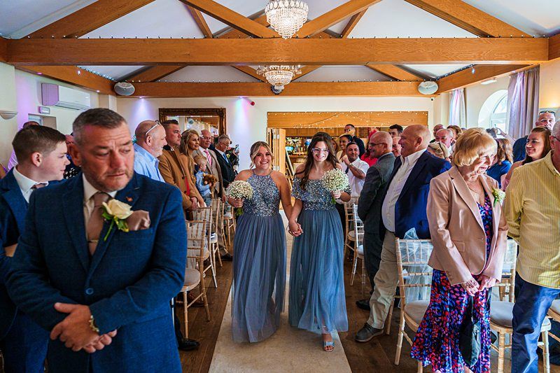 Two bridesmaids in blue dresses holding white bouquets walk down the aisle of a decorated event hall, surrounded by a smiling and applauding crowd. The hall features exposed wooden beams and chandeliers, with guests seated and standing on either side of the aisle. Image by Documentary Wedding Photographer Lewis Bishop Photography.