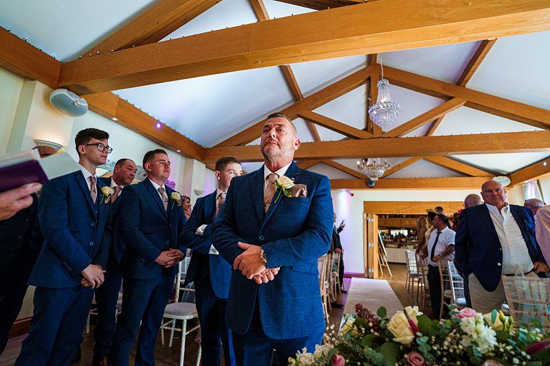 A group of groomsmen, all dressed in navy blue suits, stand in a decorated room with wooden beams and chandeliers. The groom, positioned in the center, looks ahead with anticipation. Guests are seated in the background, and floral arrangements adorn the space. Image by Documentary Wedding Photographer Lewis Bishop Photography.