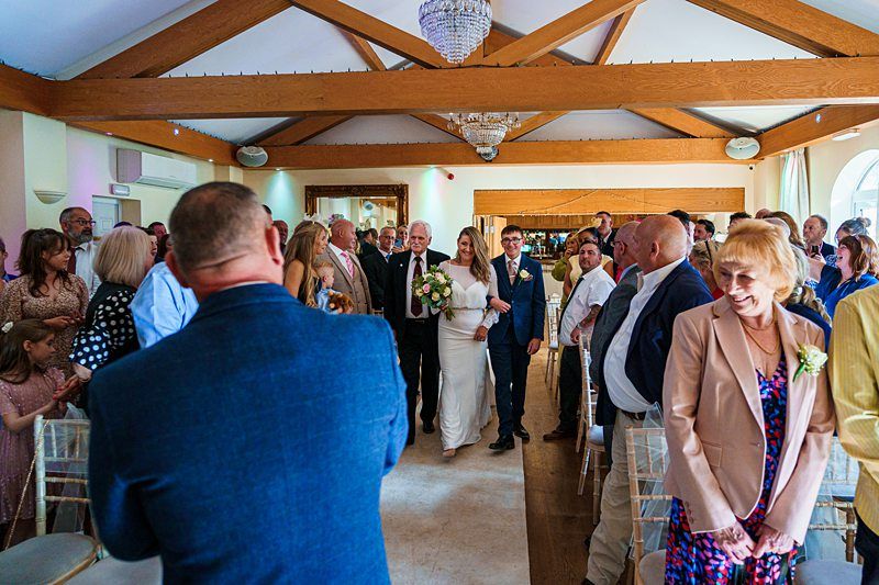 A bride in white walks down the aisle with an older gentleman, possibly her father, while guests stand and watch. The groom waits at the end of the aisle. The venue features wooden beams, chandeliers, and natural light filtering in through large windows. Image by Documentary Wedding Photographer Lewis Bishop Photography.