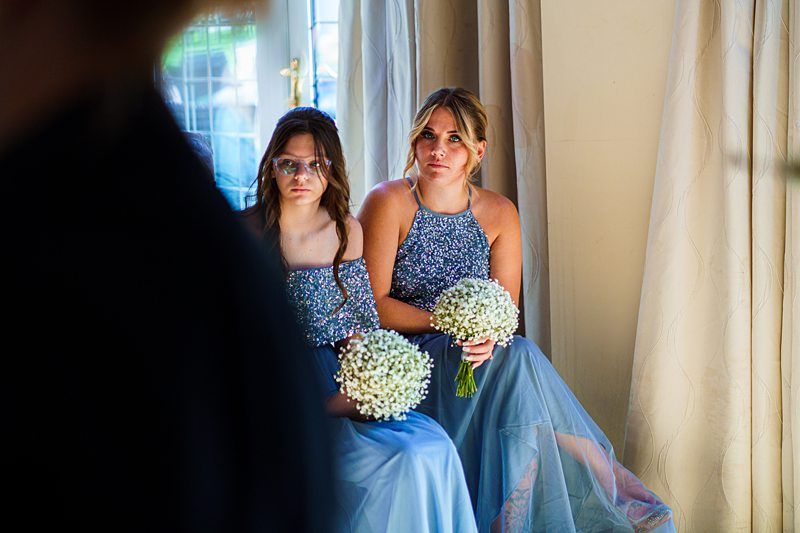 Two women in blue sequin dresses sit indoors holding bouquets of white flowers, likely as part of a wedding party. They look toward something off-camera, with natural light streaming through a window and curtains in the background. Image by Documentary Wedding Photographer Lewis Bishop Photography.