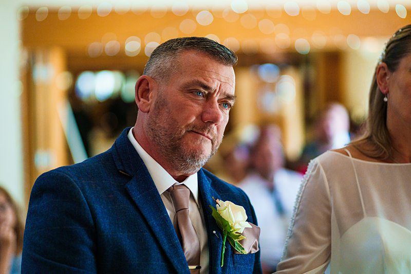 A man with short gray hair and a beard is wearing a blue suit jacket, white shirt, and beige tie, with a flower boutonnière pinned to his lapel. He is standing indoors with blurred people and lights in the background. A woman next to him is partially visible. Image by Documentary Wedding Photographer Lewis Bishop Photography.