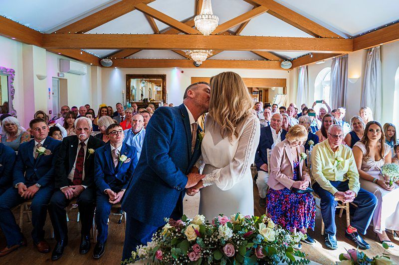 A couple, dressed in wedding attire, kiss at the altar in a room filled with guests. They stand behind a floral arrangement, while guests, seated in rows, watch the ceremony. The room is brightly lit with wooden beams and chandeliers overhead. Image by Documentary Wedding Photographer Lewis Bishop Photography.