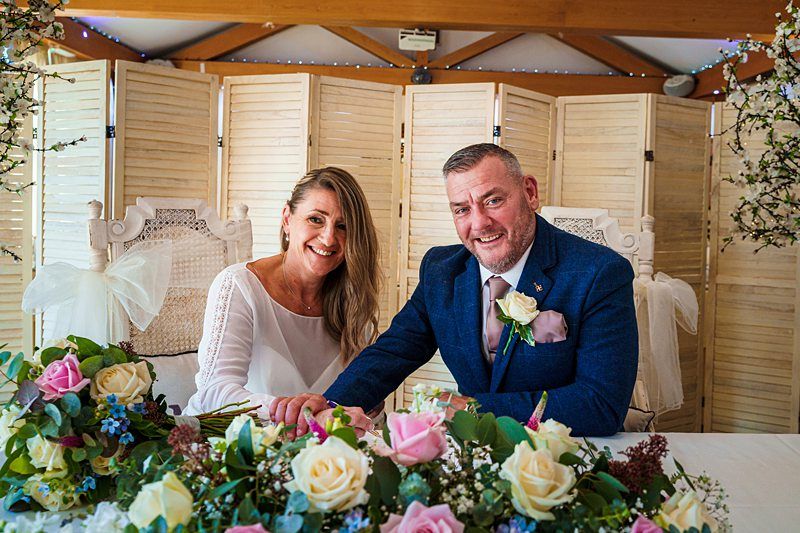 A smiling couple sits behind a table adorned with colorful flowers. The woman, with long blonde hair, wears a white dress, and the man, dressed in a blue suit, sports a tie. They hold hands and pose in front of a wooden screen decorated with fairy lights. Image by Documentary Wedding Photographer Lewis Bishop Photography.
