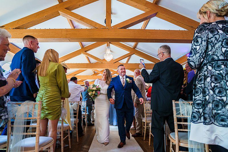 A newlywed couple walks down the aisle, smiling and holding hands. The groom, in a blue suit, and the bride, in a white dress, are surrounded by applauding guests. The venue has wooden beams and chandeliers overhead. A man is taking their photo with a smartphone. Image by Documentary Wedding Photographer Lewis Bishop Photography.