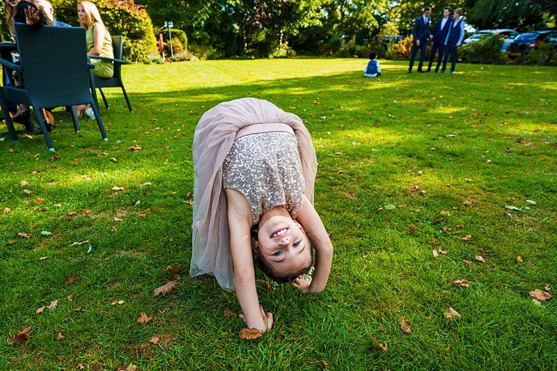 A young girl in a sparkling dress is doing a backbend on a grassy lawn. She is smiling at the camera, and there are adults sitting at a table in the background, along with a few others standing and walking on the lawn. The weather is sunny with green trees all around. Image by Documentary Wedding Photographer Lewis Bishop Photography.