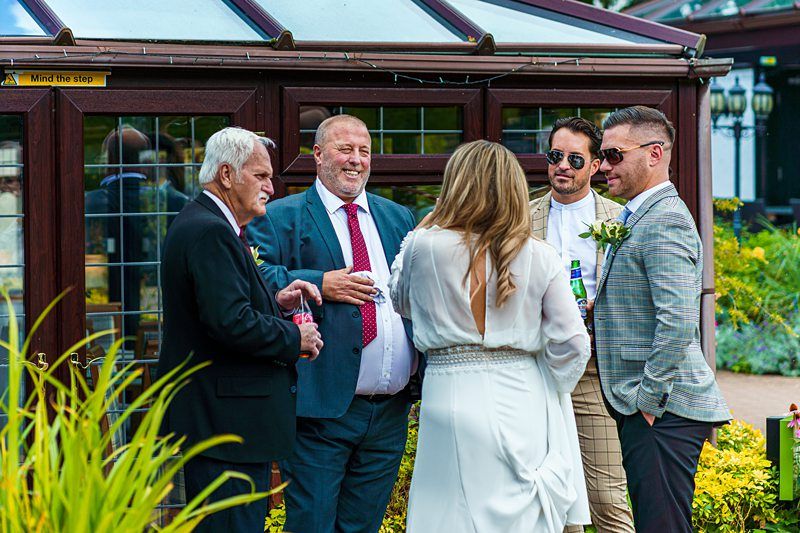 A small group of people is gathered outside near a building with large windows. Three men and a woman are engaged in conversation. The men are wearing suits, while the woman in the center is wearing a white dress and has her back to the camera. Image by Documentary Wedding Photographer Lewis Bishop Photography.
