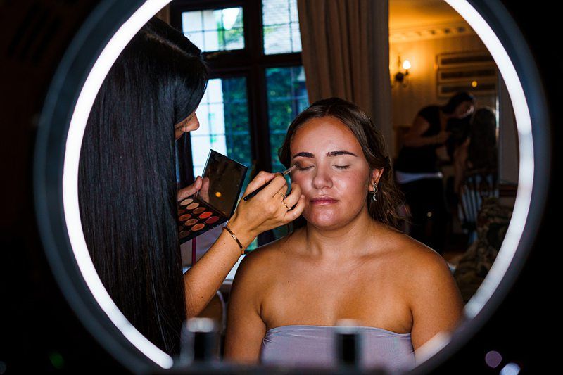 A makeup artist applies eyeshadow to a woman with her eyes closed, illuminated by a bright circular ring light in the foreground. The woman is wearing a strapless top and appears relaxed. The background shows a room with more people and ambient lighting. Image by Documentary Wedding Photographer Lewis Bishop Photography.